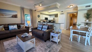 A living room with a brown sofa, a coffee table, and a dining area with a white table and chairs. - Newly Renovated Beachfront w/ Coastal Style! Free Golf & Daily Activities! (Panama City Beach)