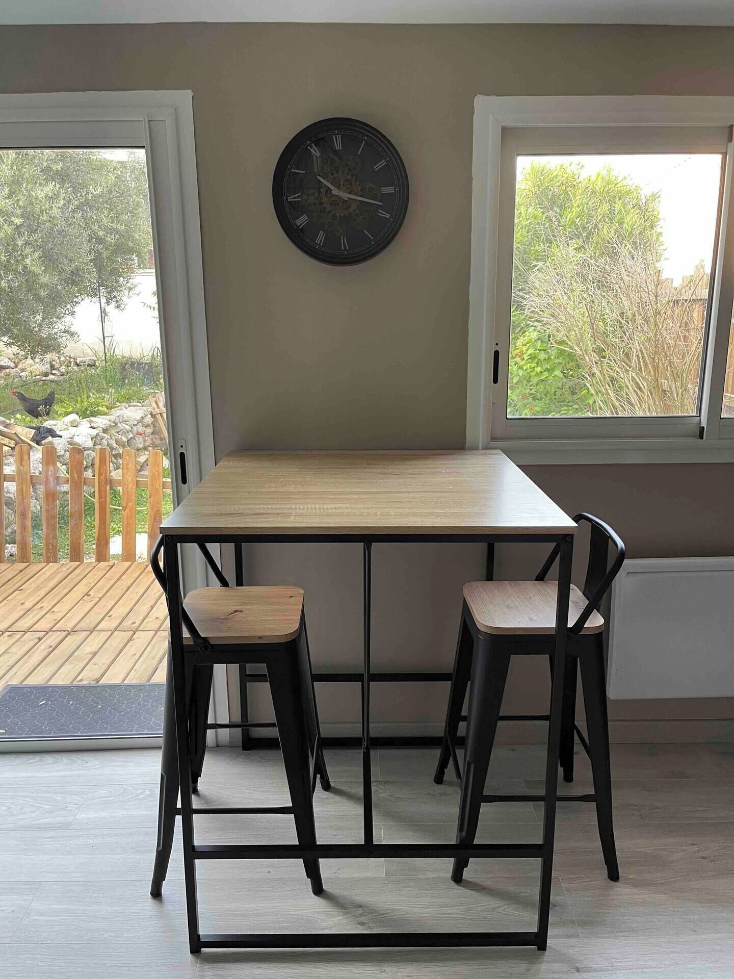 Un coin repas avec une table en bois et deux chaises, une horloge murale et une vue sur un jardin par la fenêtre.