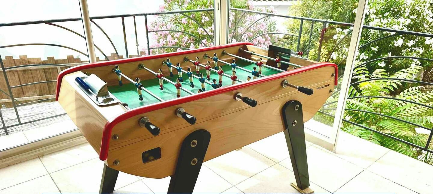 A baby-foot foosball table with a wooden base and red and black handles, placed on a balcony with a view of greenery and a wooden fence.