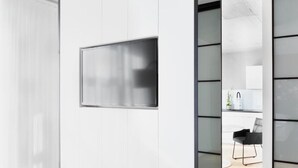 Ein modernes Interieur mit einem großen Flachbildfernseher an einer weißen Wand, einer Küchenzeile mit weißen Schränken und einem schwarzen Stuhl sowie einem Holzboden.