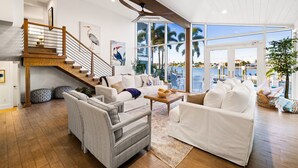 A spacious living room with a wooden staircase, white sofas, and large windows offering a view of the ocean.