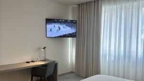 A hotel room with a bed, a desk, a chair, a television displaying a hockey game, and a window with curtains.