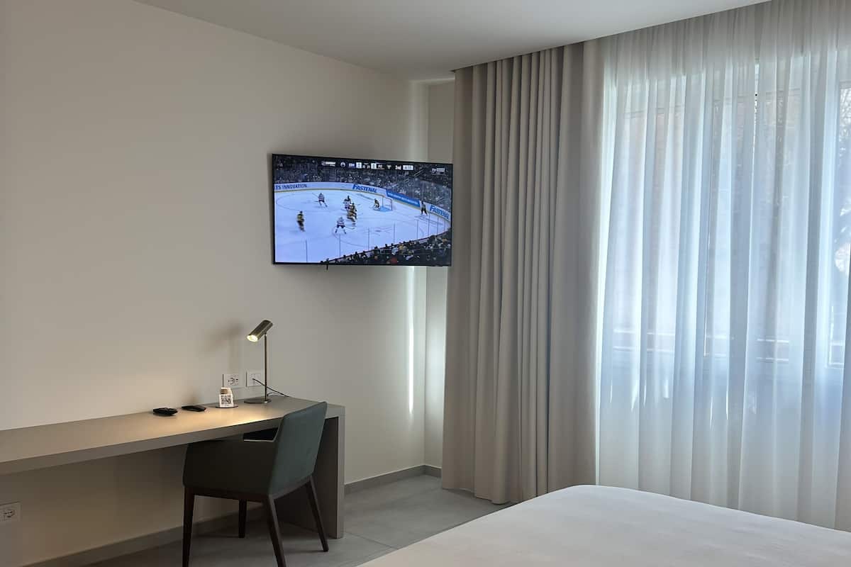 A hotel room with a bed, a desk, a chair, a television displaying a hockey game, and a window with curtains.