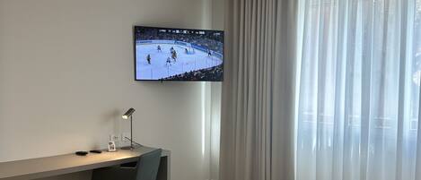Habitación de hotel con cama, escritorio, silla, televisor mostrando un partido de hockey y ventana con cortinas.