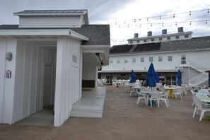 A covered outdoor seating area with white plastic chairs and tables, a women's restroom sign, and a building with a sloped roof in the background.