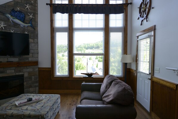 A living room with a fireplace, a television mounted on a stone wall, a window with blinds, a wooden door, and a view of the outside.