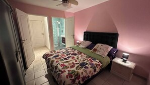 Un dormitorio con cama, mesitas de noche, ventilador de techo y puerta que da a un baño.