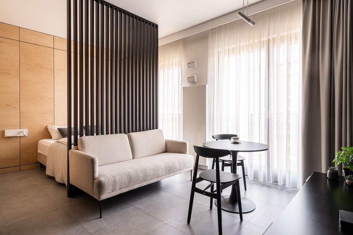 Uma sala de estar moderna com um sofá, uma mesa de jantar e cadeiras. Há uma janela com cortinas, uma planta e um luminário fixado na parede.