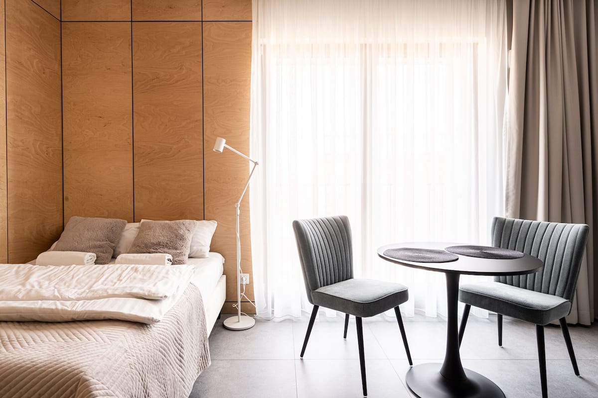 Quarto de hotel moderno com parede de painel de madeira, cama com roupa de cama bege, luminária de chão e área de jantar com duas cadeiras e uma mesa redonda.