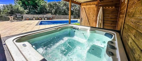 A hot tub with a wooden deck and a pool in the background.