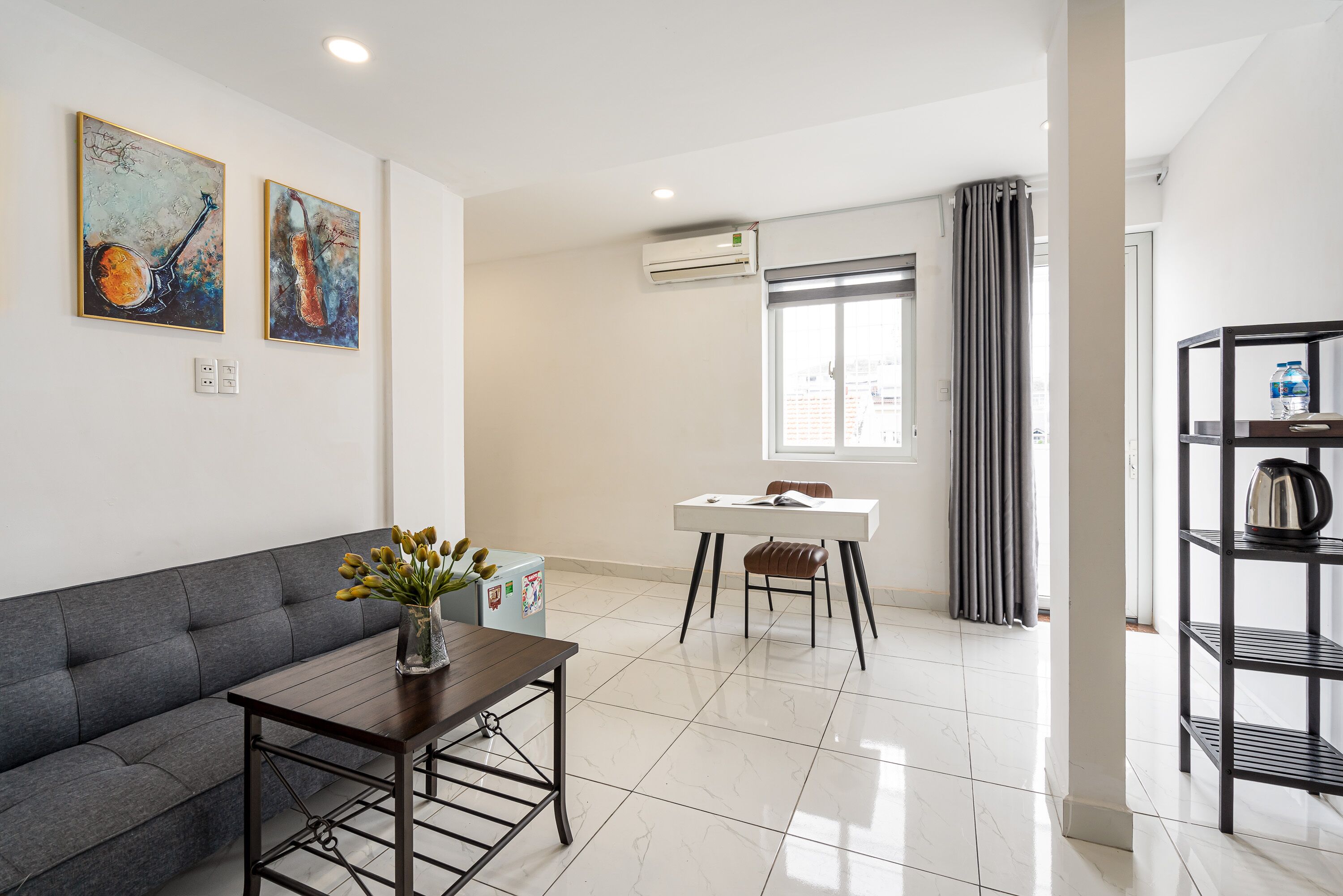 A modern living room with a grey sofa, a wooden coffee table, and a dining area with a white table and chairs. There are two paintings on the wall, an air conditioning unit, and a water dispenser on a shelf.