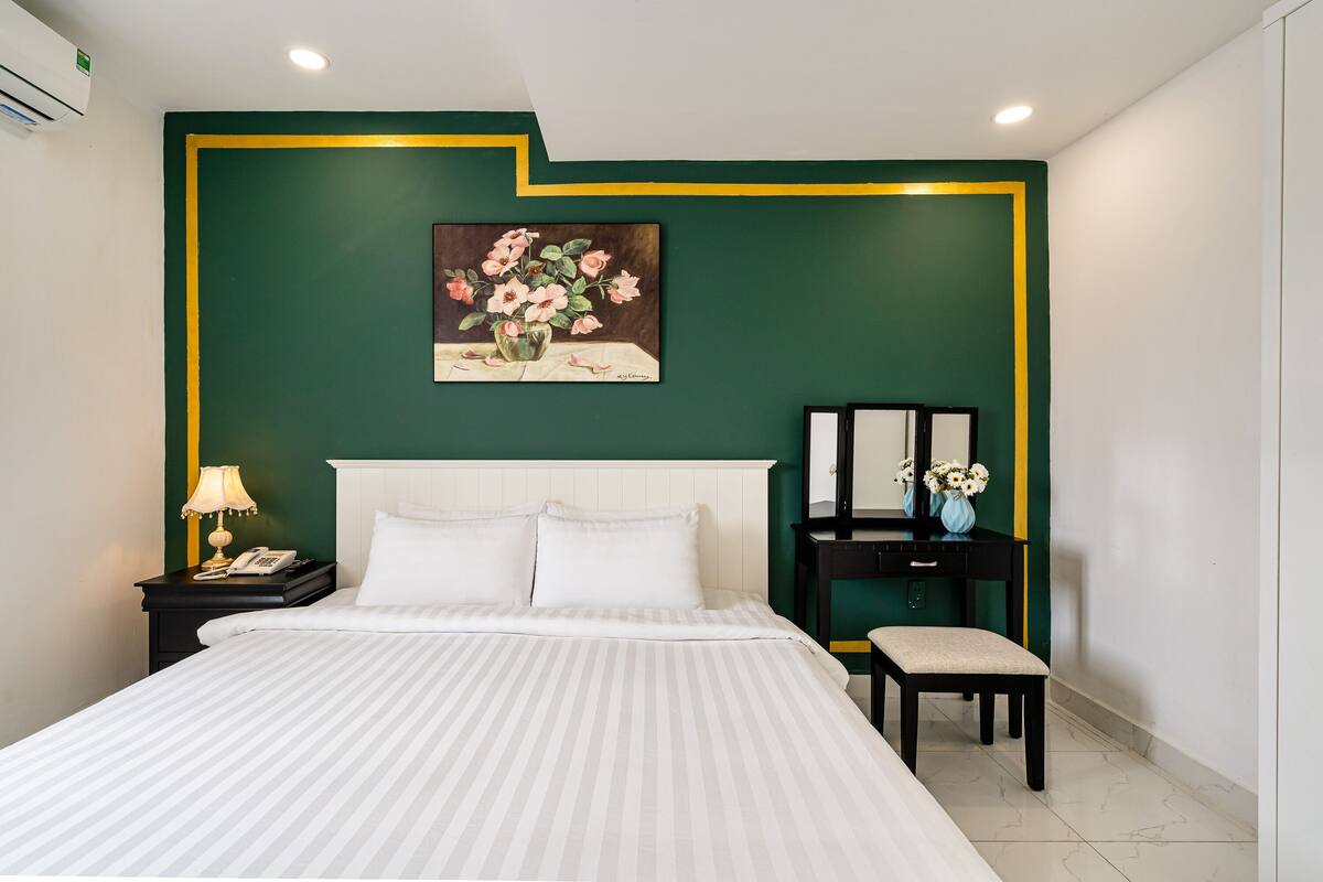 Une chambre d’hôtel avec un grand lit, une table de chevet avec une lampe, une commode, une coiffeuse, un banc et un tableau représentant des fleurs accroché au mur.