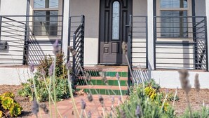 Une maison avec un porche à l’avant, une porte noire et un petit jardin agrémenté de plantes et d’un chemin en briques.