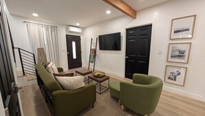 Un salon moderne avec un canapé, un fauteuil et une table basse. Un téléviseur est fixé au mur et une porte noire est visible.
