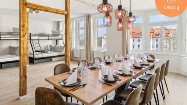 Een eetruimte met een lange houten tafel gedekt voor zes personen, omringd door stoelen. Boven de tafel hangen hanglampen.
