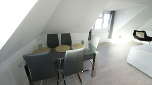 Ein modernes Schlafzimmer im Loft-Stil mit schrägem Dach, einem Glastisch mit Stühlen, einem weißen Sofa und einem großen Fenster, das für natürliches Licht sorgt.