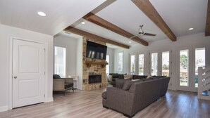 A spacious living room with a stone fireplace, wooden beams, and large windows. - Changes in Latitude by Lake Martin Vibe (Dadeville)