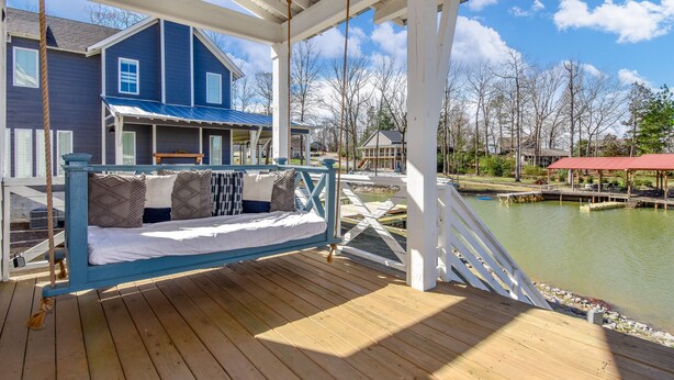 A wooden deck with a swing chair overlooking a body of water, a blue house, and a white gazebo. - Changes in Latitude by Lake Martin Vibe (Dadeville)