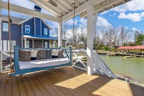A wooden deck with a swing chair overlooking a body of water, a blue house, and a white gazebo. - Changes in Latitude by Lake Martin Vibe (Dadeville)