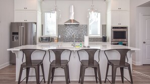 A modern kitchen with white cabinetry, a central island with bar stools, stainless steel appliances, and pendant lighting. - Changes in Latitude by Lake Martin Vibe (Dadeville)