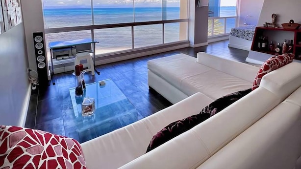 Un soggiorno moderno con una grande finestra vista mare, un divano bianco, un tavolino in vetro e un piccolo tavolino con una stampante.