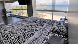 Una camera da letto con copriletto a motivi zebrati, un'ampia finestra vista spiaggia e una televisione a schermo piatto montata a parete.