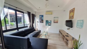A modern living room with a black sofa, a glass coffee table, a wooden TV stand, and a large window with curtains. - D&A Paradise Villas (Koh Samui)