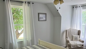 A bedroom with a bed, a chair, a ceiling fan, and a window with curtains. - The Modern Maine Retreat (Oakland)