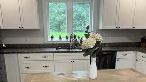A kitchen with white cabinets, a granite countertop, a sink, and a vase with flowers. There are two stools and a window with a view of trees. - The Modern Maine Retreat (Oakland)