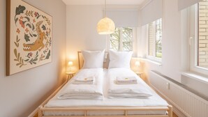 Un lit double avec du linge de lit blanc, deux tables de chevet avec des lampes, un luminaire suspendu, une grande œuvre d’art florale accrochée au mur et une fenêtre donnant sur un paysage verdoyant.