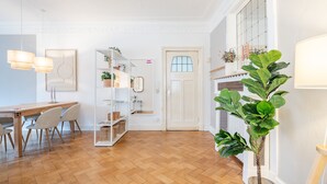 Un intérieur moderne avec un plancher en bois, un coin repas comprenant des chaises et une table, une étagère blanche ornée de plantes et une grande plante en pot.