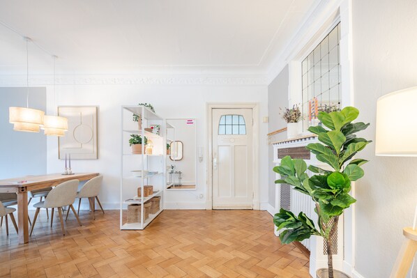 Een modern interieur met een houten vloer, een eethoek met stoelen en een tafel, een witte boekenkast met planten, en een grote potplant.