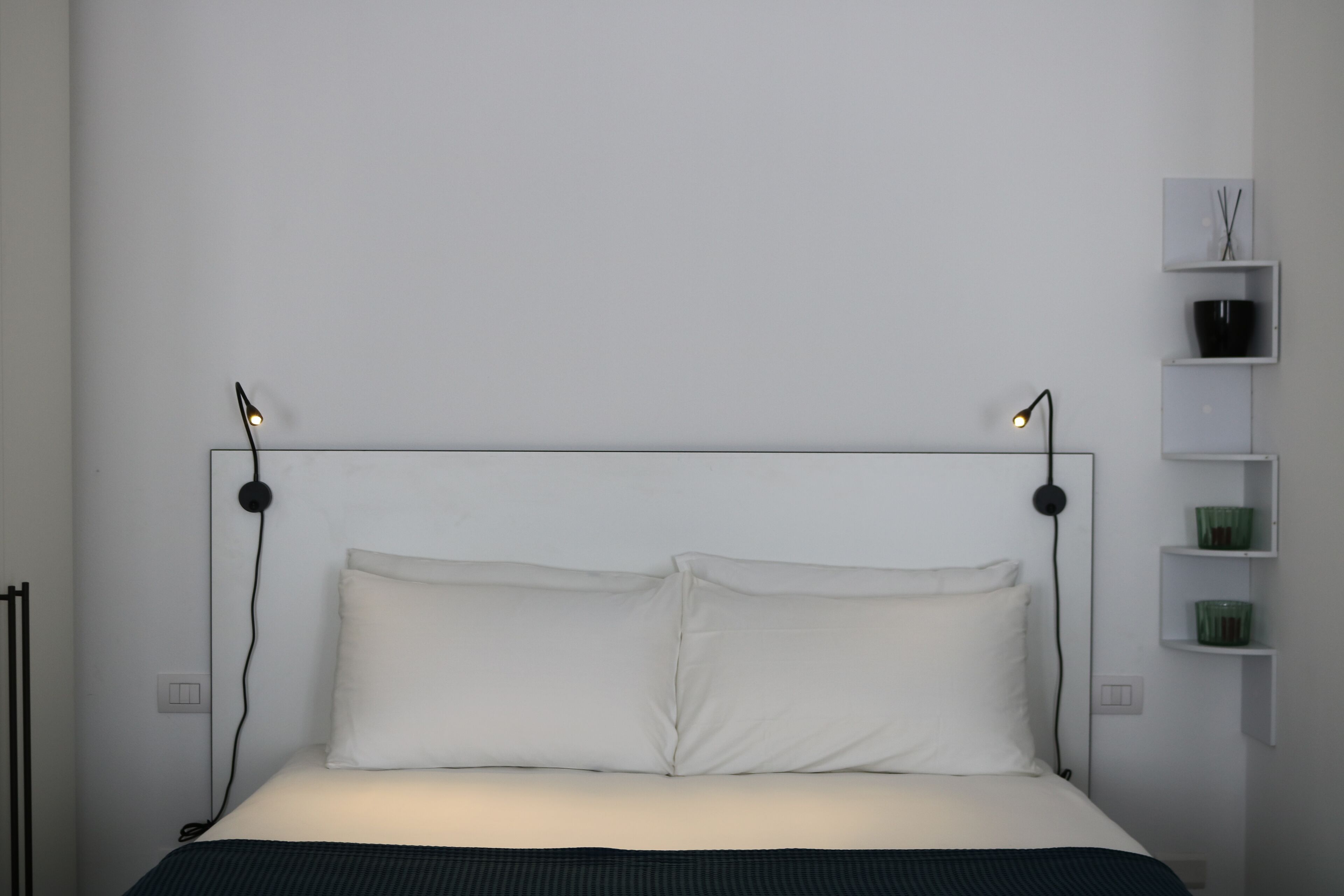 A modern bedroom with a bed, bedside lamps, and a white wall with shelves.