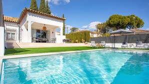 Une piscine à l’eau d’un bleu limpide, entourée d’un bâtiment blanc doté d’un toit en tuiles et d’un espace extérieur aménagé et couvert pour s’asseoir.