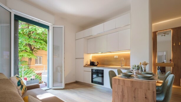 A modern kitchen with white cabinets, a wooden dining table, and a sliding glass door leading to a balcony with plants.