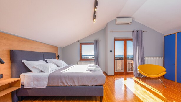A modern bedroom with a large bed, a wooden headboard, a bright yellow chair, and a view of the outdoors through a sliding door.