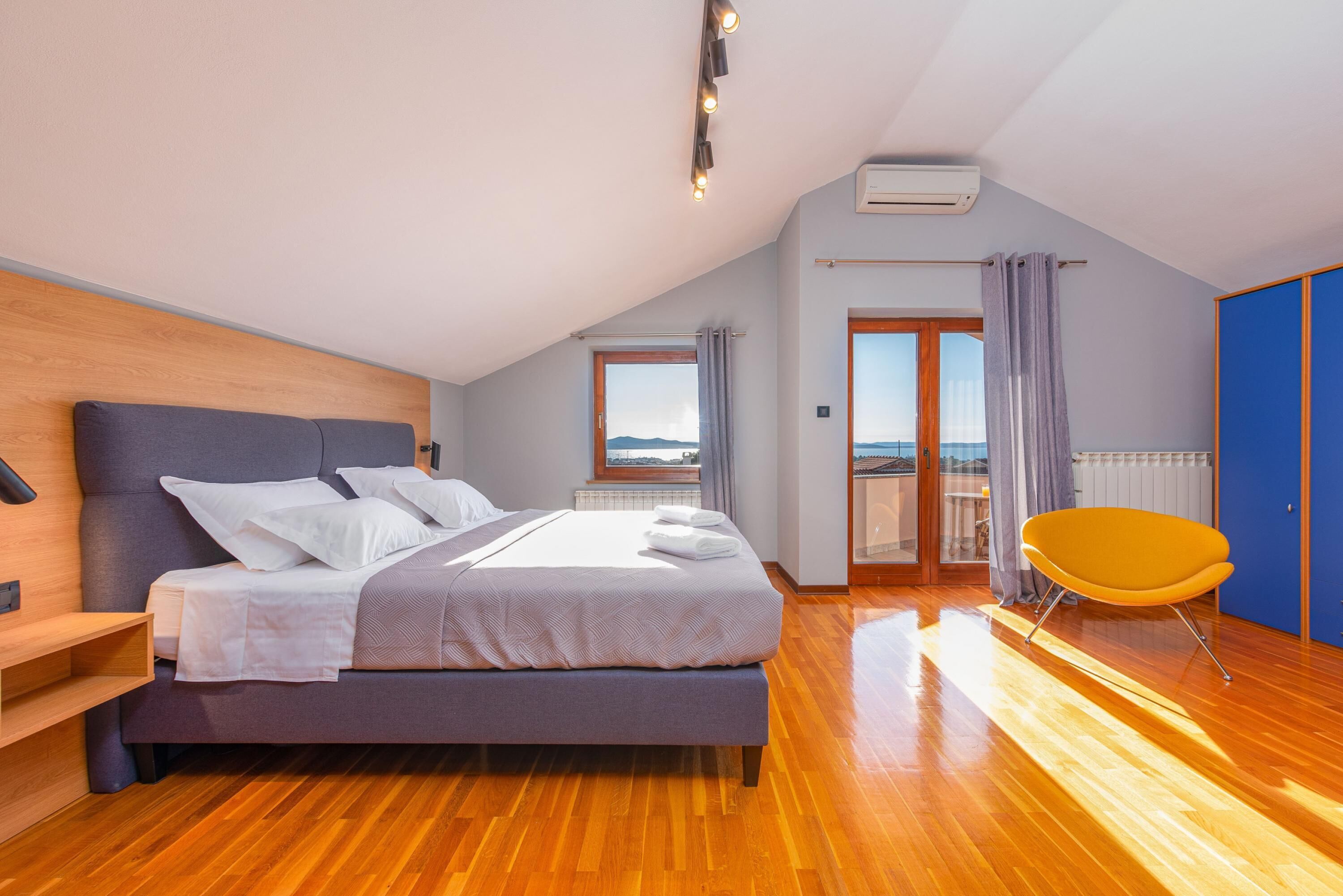 Ein modernes Schlafzimmer mit einem Holz-Kopfteil, einem großen Bett mit weißer Bettwäsche, einem leuchtend gelben Sessel und Blick ins Freie durch ein großes Fenster.