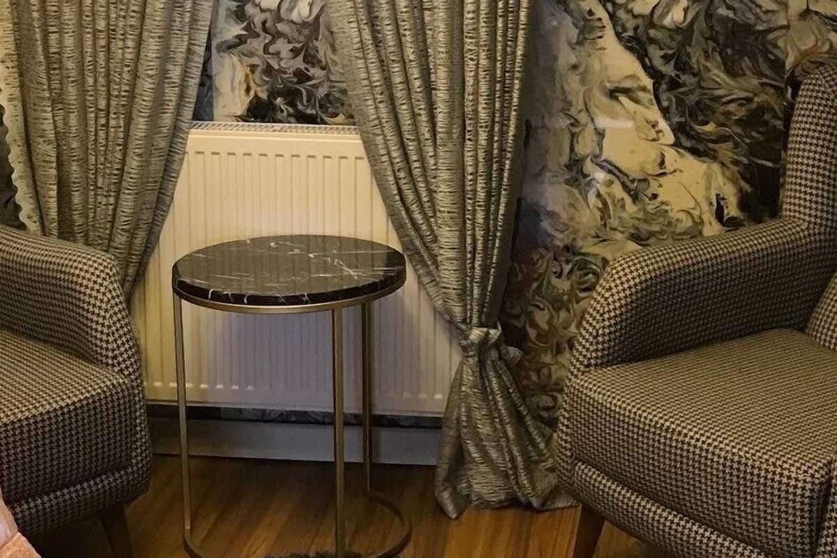 A room with a radiator, two patterned chairs, a small round table, and curtains with a floral design.