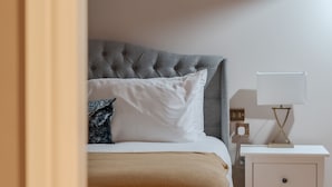 Un lit bien fait, doté d’une tête de lit capitonnée grise, de coussins blancs à motifs et d’un plaid beige. On aperçoit une table de chevet avec une lampe blanche et une petite table de chevet avec un cadre doré et une lampe blanche.