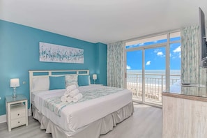 A bedroom with a bed, a nightstand, a lamp, a painting, and a view of the beach.