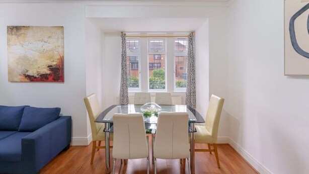 A modern living room with a glass dining table, white chairs, a blue sofa, and a large abstract painting on the wall. - British Character in Golders Green. ground floor. 20min to oxford st. (London)