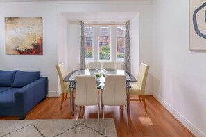 A modern living room with a glass dining table, white chairs, a blue sofa, and a large abstract painting on the wall.
