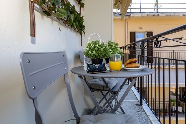 Un balcon avec une petite table, un pot de fleurs, un verre de jus d’orange et une assiette de croissants.