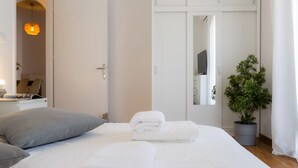 Una cama bien hecha con sábanas blancas, una almohada gris y una planta en maceta al lado de un armario blanco.
