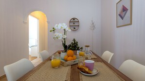 Una mesa de comedor con un jarrón de orquídeas blancas, una bandeja de naranjas, un vaso de zumo de naranja, un croissant y una taza rosa.