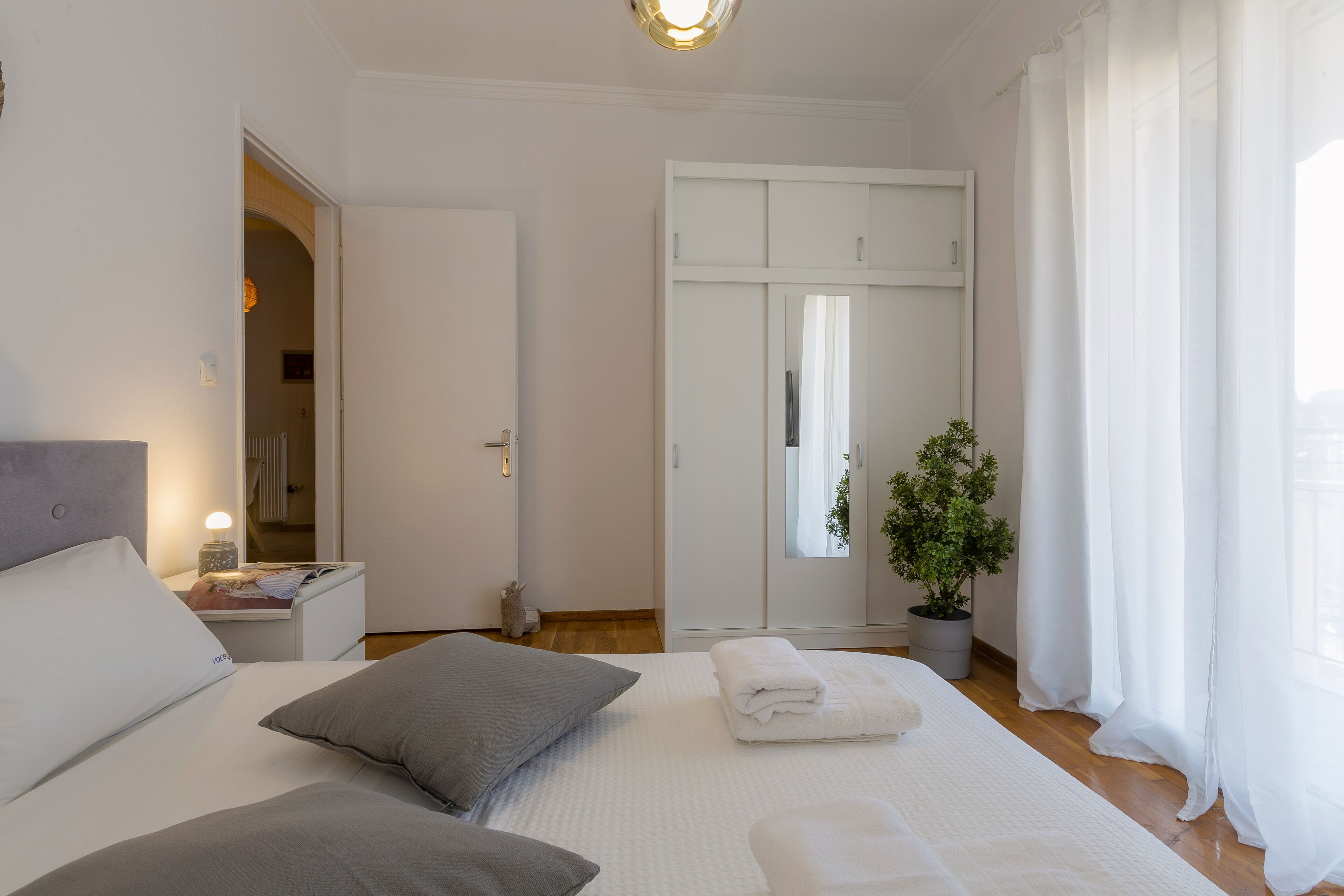 Un dormitorio con cama, almohadas, una mesita de noche, un armario, un espejo, una planta en maceta y una ventana con cortinas.