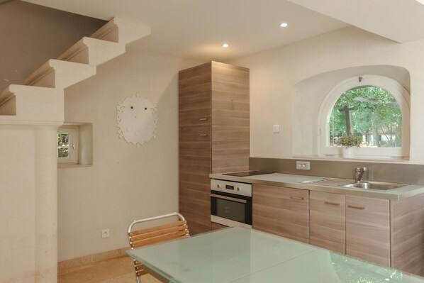 A modern kitchen with a glass dining table, wooden cabinets, and a built-in oven. - Gîte D'annette au Château des Barrenques (Lamotte-du-Rhône)