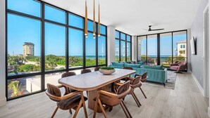 A modern dining area with a large table and chairs, large windows offering a view of the sea and buildings, and a ceiling fan.