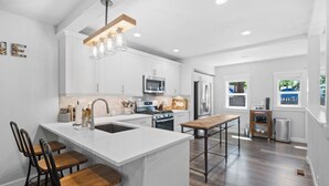 Una cocina moderna con armarios blancos, una isla central con taburetes de bar, electrodomésticos de acero inoxidable y una lámpara colgante.