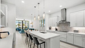 A modern kitchen with a central island, bar stools, and a view to a living area. - Ocean's Edge | Lavish NEW Condo w Beach Views, Cozy Decor & Community Amenities (Melbourne Beach)